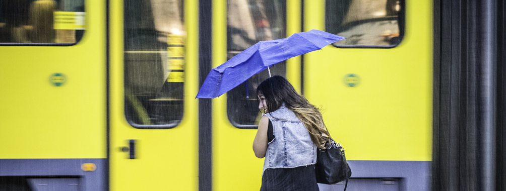 Junge Frau mit blauem Regenschirm vor gelber Straßenbahn