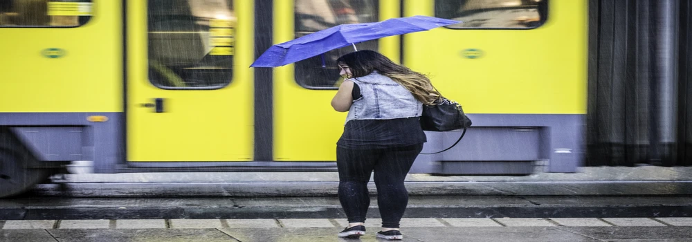 Junge Frau mit blauen Regenschirm vor gelber Stadtbahn