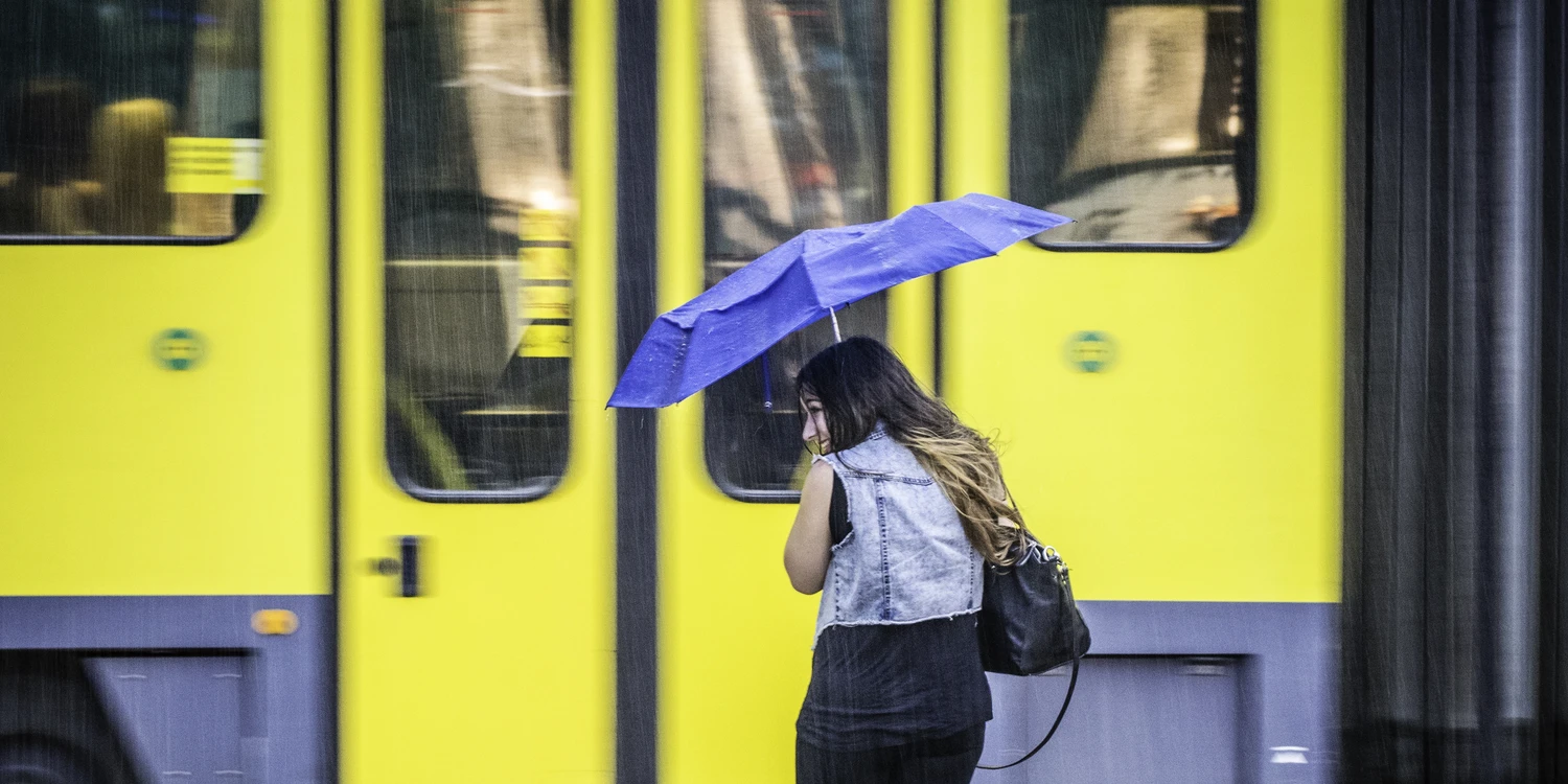 Junge Frau mit blauen Regenschirm vor gelber Stadtbahn