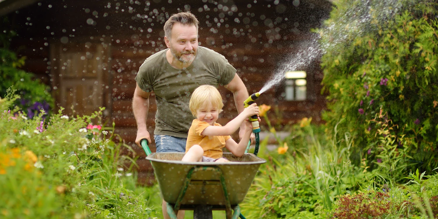 Kleiner Junge mit Gartenschlauch wird von seinem Vater in einer Schubkarre geschoben