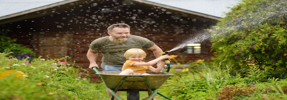 Kleiner Junge mit Gartenschlauch wird von seinem Vater in einer Schubkarre geschoben